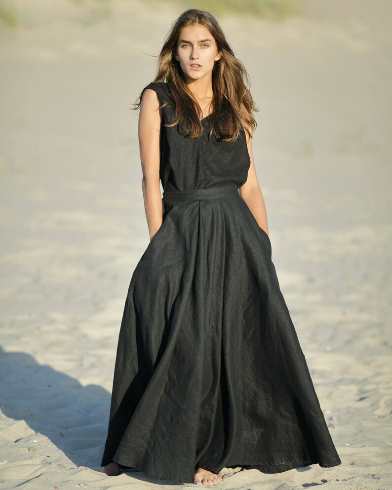 May include: A sleeveless black dress with a V-neck and a flowing, floor-length skirt. The dress has a fitted waist and pockets. The model is standing on a sandy beach.