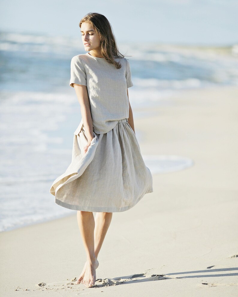 K&ouml;nnte beinhalten: Ein hellbeige Leinen-Zweiteiler, bestehend aus einem kurz&auml;rmeligen Oberteil und einem knielangen Rock. Das Outfit wird an einem Sandstrand mit dem Meer im Hintergrund getragen. Der Rock ist leicht ausgestellt und der Stoff wirkt leicht und flie&szlig;end.