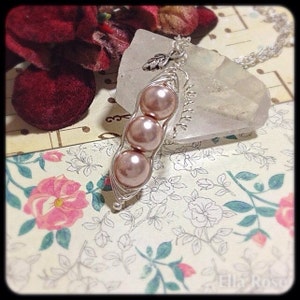 May include: A silver necklace with a charm that looks like a pea pod with three pink pearls inside. The charm is hanging from a clear crystal pendant.