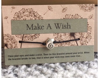 May include: A black string bracelet with a silver seashell charm. The card reads "Make A Wish" and includes instructions to tie the bracelet around your wrist and make a wish. When the bracelet breaks, your wish may come true.