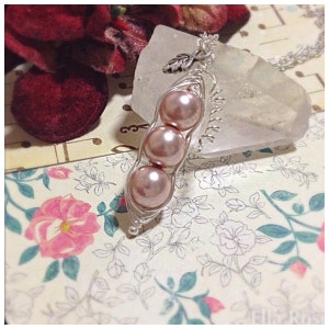 May include: A silver necklace with a charm of three pink pearls in a pea pod design. The necklace is on a clear crystal surface.