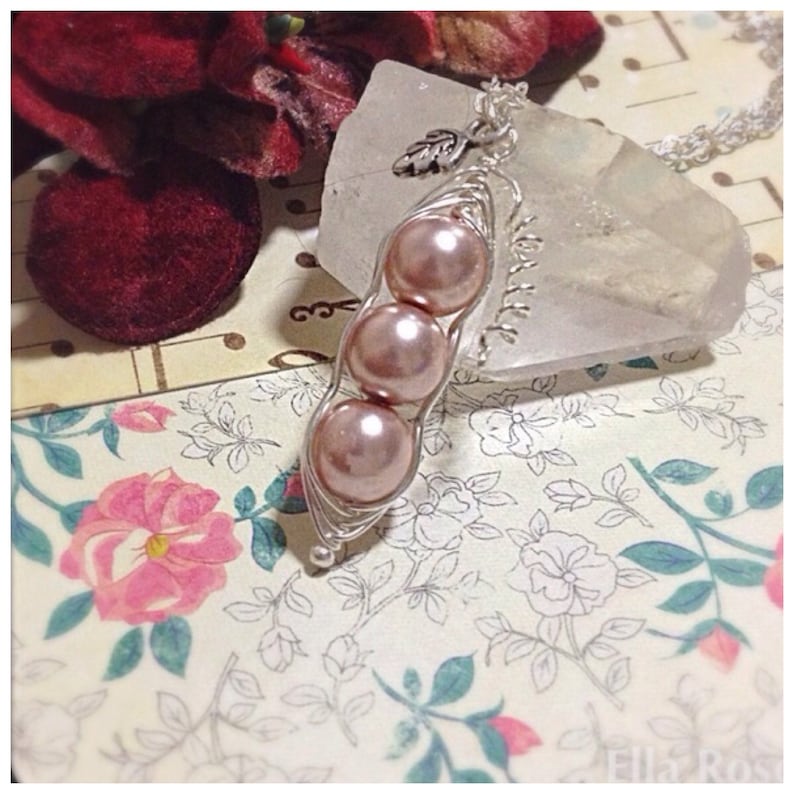 May include: A silver necklace with a charm of three pink pearls in a pea pod design. The necklace is on a clear crystal surface.