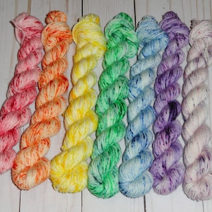 May include: A selection of eight skeins of colourful yarn arranged in a rainbow sequence. The yarn colours include pink, orange, yellow, green, blue, purple, and white. Each skein is tightly wound and speckled with contrasting colours.