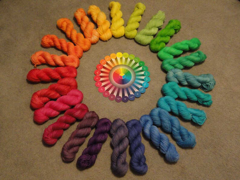 May include: A rainbow color wheel with yarn skeins arranged in a circle around it. The yarn skeins are in different shades of red, orange, yellow, green, blue, indigo, and violet.