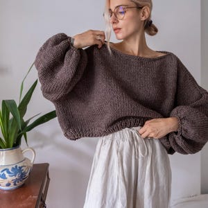 May include: A brown knit sweater with voluminous sleeves. The sweater has a wide neckline and a cropped fit. The model is wearing the sweater with light-colored linen pants. The sweater is a warm, neutral color, suitable for fall or winter.