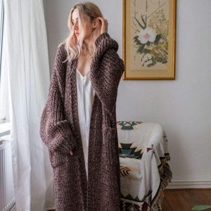 May include: A long, burgundy knit cardigan with large pockets and a hooded design. The oversized sweater is worn over a white dress. The cardigan is a chunky knit with a textured appearance.
