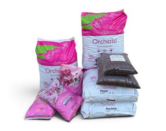 May include: A collection of Orchiata orchid bark potting medium bags in various sizes. The packaging is white and pink with orchid flower graphics and text including Orchiata, Power, and Precision. Some bags are clear plastic showing dark bark.