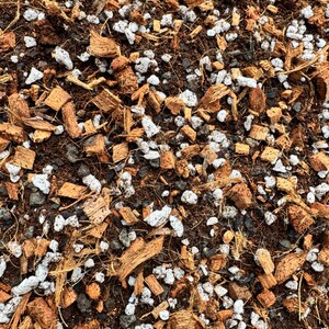 May include: Close-up of a mix of potting soil components. The image shows a blend of dark brown soil, light brown coconut husk pieces, white perlite, and small gray rocks, creating a textured, natural appearance.