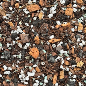 May include: Close-up of a mix of potting soil components. The blend includes dark soil, light-colored perlite, brown bark chips, and small pieces of coconut husk. The texture is coarse and varied, with a natural, earthy appearance.