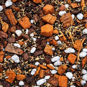 May include: Close-up of a soil mix for plants, featuring a blend of brown, orange, and white components. The mix includes pieces of bark, small white pebbles, and dark soil, creating a textured, natural appearance.