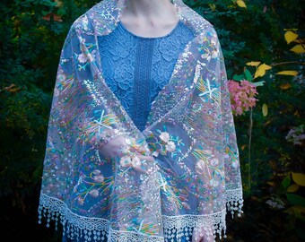 May include: A blue lace dress and a sheer shawl with floral embroidery and sequin accents. The shawl has a white lace trim with dangling beads. The background is a lush green garden with pink flowers.