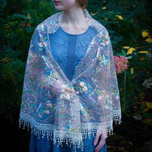 May include: A blue lace dress and a sheer shawl with floral embroidery and sequin accents. The shawl has a white lace trim with dangling beads. The background is a lush green garden with pink flowers.