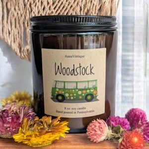 Puede incluir: Una vela en tarro de cristal marrón con tapa negra. La etiqueta dice "Woodstock" con una ilustración de autobús verde. La vela es de 227g y vertida a mano en Pensilvania. Flores secas amarillas, rosas y moradas rodean la vela.