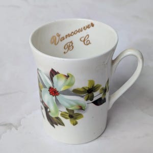 May include: White ceramic mug with a floral design featuring light green and yellow flowers with a red center. The mug has a curved handle and the words "Vancouver B.C." in gold lettering.