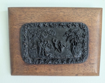 May include: A rectangular wooden plaque with a dark bronze-colored relief. The relief depicts a detailed scene with figures, trees, and architectural elements. The plaque is framed by a darker wood.