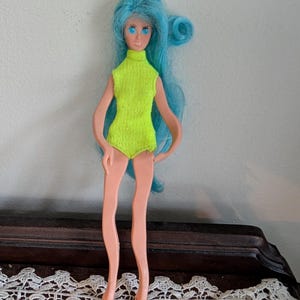 May include: A vintage doll with long, light blue hair and blue eyes. The doll is wearing a neon green sleeveless bodysuit. The doll is standing on a dark brown surface with a white lace trim.