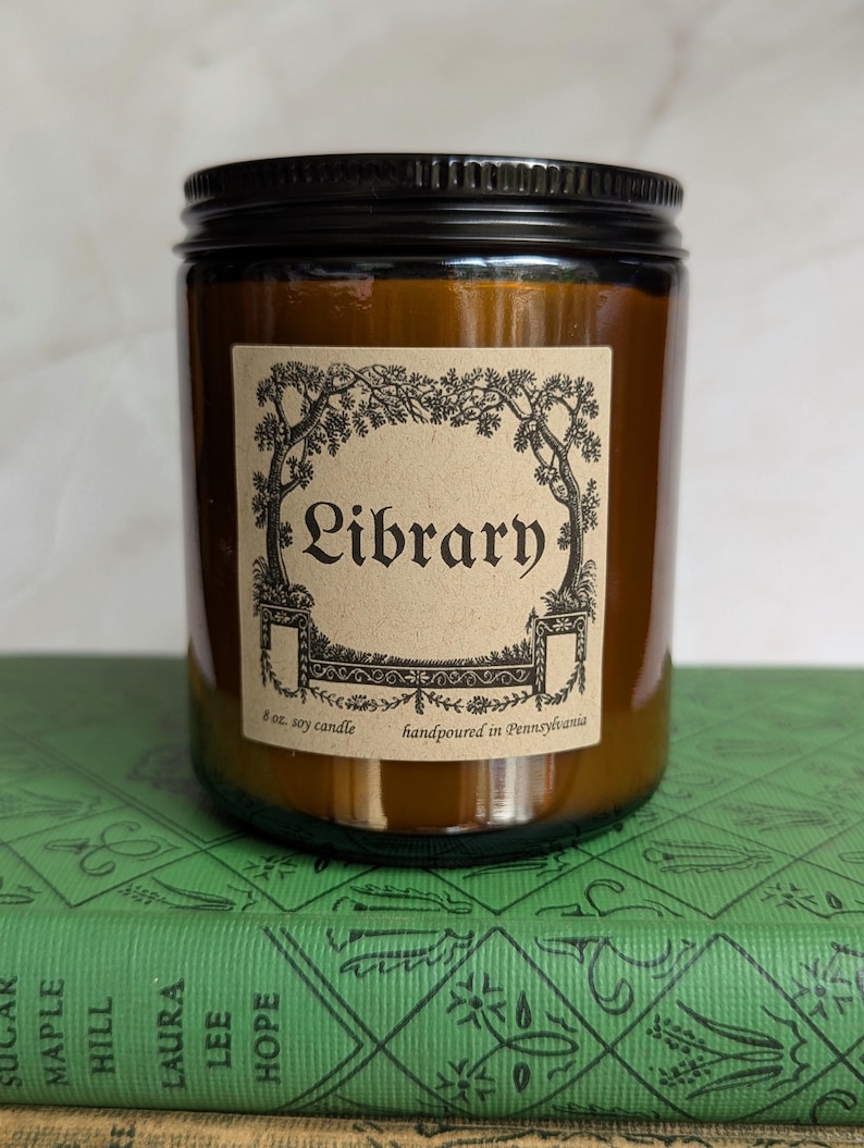 May include: An 8 oz. soy candle in an amber glass jar with a black metal lid. The beige rectangular label features a black ink illustration of trees and the text Library, 8 oz. soy candle, and handpoured in Pennsylvania.