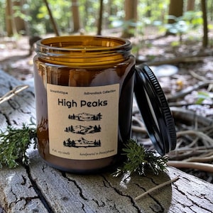 May include: A brown glass jar candle with a black lid, labeled "High Peaks" from the "Adirondack Collection." The label features mountain illustrations. The candle is 8 oz and handpoured in Pennsylvania. The jar sits on a weathered wooden surface.