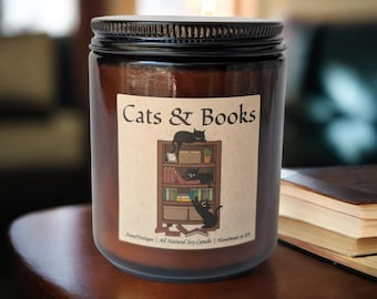 Könnte beinhalten: Eine braune Glas-Kerze mit schwarzem Deckel und brennendem Docht. Das Etikett zeigt den Text Cats & Books über einer Illustration eines hölzernen Bücherregals mit Büchern und drei schwarzen Katzen. Der untere Text lautet AnnaVintique | All Natural Soy Candle | Handmade in US.