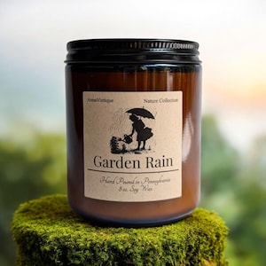 May include: A brown glass jar candle with a black lid, labeled "Garden Rain." The label features a silhouette of a person watering plants under an umbrella. The candle is 8 oz. and made with soy wax. The candle sits on a mossy surface.