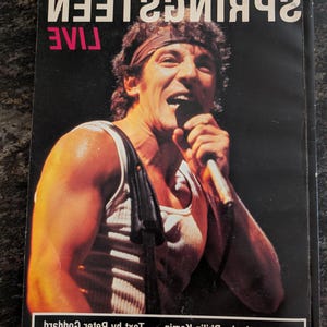 Puede incluir: Una portada de álbum de concierto con Bruce Springsteen cantando a un micrófono. El título del álbum "SPRINGSTEEN LIVE" se muestra en blanco, con "LIVE" en rosa. La portada también incluye el texto "fotografías de Kamin, texto de Goddard."