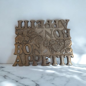 Op de afbeelding: Een bronzen metalen bord met de woorden "BON APPETIT" in vette hoofdletters. Het bord bevat illustraties van aardbeienplanten tussen de woorden. Het bord is rechthoekig met een licht getextureerd oppervlak.