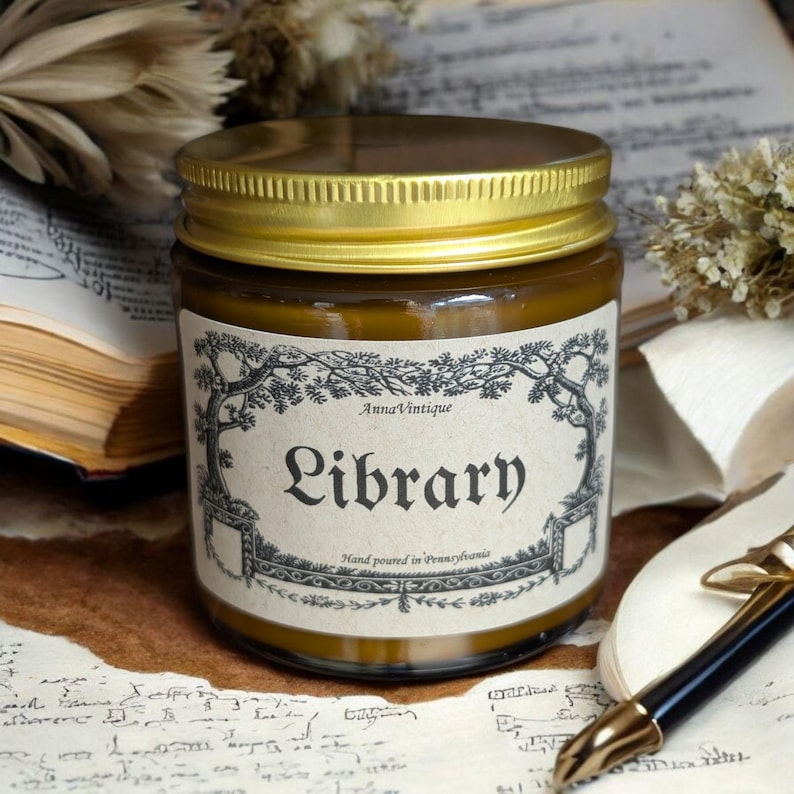 May include: A glass candle jar with a gold-colored metal lid and a tan label featuring black decorative borders and the text AnnaVintique, Library, and Hand poured in Pennsylvania. The candle wax inside the jar is a dark amber brown color.