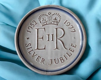 May include: A round, off-white ceramic plate with a blue rim. The plate features the text "1952 SILVER JUBILEE 1977" and the initials "ER II" beneath a crown design. The plate is set against a blue background.