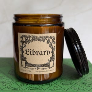 May include: An amber glass jar candle with a black metal lid. The label features a decorative black ink illustration of trees and the text Library, 8 oz. soy candle, handpoured in Pennsylvania. The candle is placed on a green book cover.