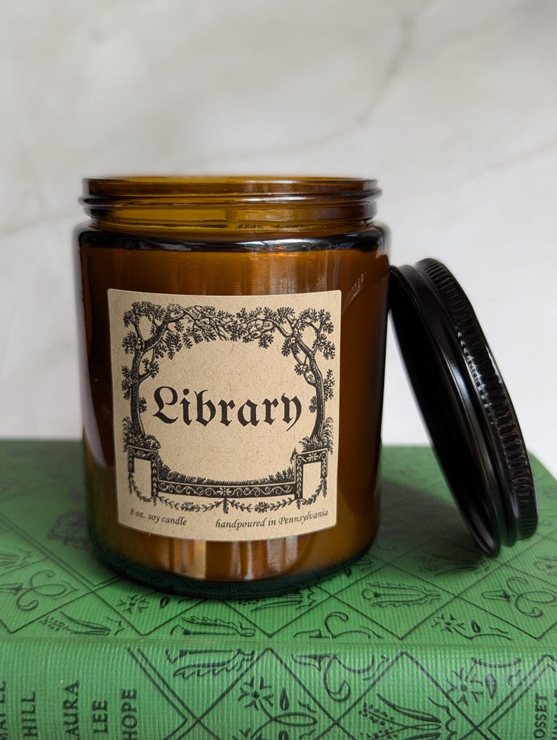 May include: An amber glass jar candle with a black metal lid. The label features a decorative black ink illustration of trees and the text Library, 8 oz. soy candle, handpoured in Pennsylvania. The candle is placed on a green book cover.