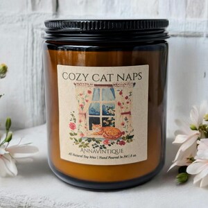 May include: A brown glass jar candle with a black lid. The label reads "COZY CAT NAPS" with an illustration of a cat by a window. The candle is made with all-natural soy wax and hand-poured in PA, with a net weight of 8 oz.