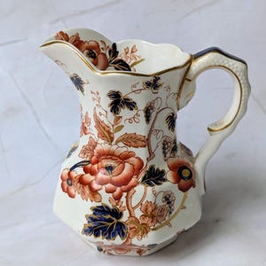 May include: A cream-colored ceramic pitcher featuring a detailed floral pattern in shades of burnt orange, deep blue, and gold. The vessel has a scalloped rim, a curved handle with textured detailing, and gold trim along the edges and handle.