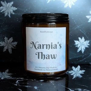 May include: A glass jar candle with a black lid and a white label. The label reads "Narnia's Thaw" in a decorative font, with "AnnaVintique" above. The candle is surrounded by snowflake decorations.