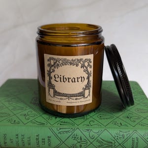 May include: An 8 oz. soy candle in an amber glass jar with a black metal lid. The label features a decorative border and the text "Library" along with "8 oz. soy candle" and "handpoured in Pennsylvania" printed in black ink on tan paper.