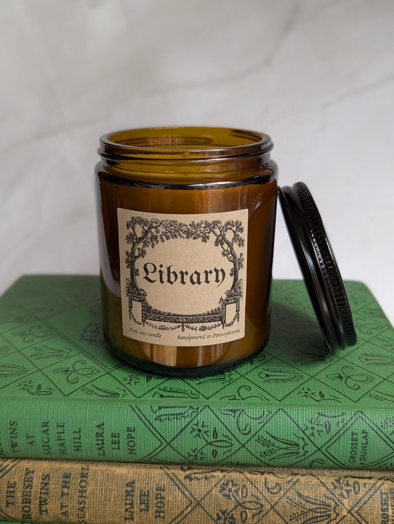 May include: An 8 oz. soy candle in an amber glass jar with a black metal lid. The label features a decorative border and the text "Library" along with "8 oz. soy candle" and "handpoured in Pennsylvania" printed in black ink on tan paper.