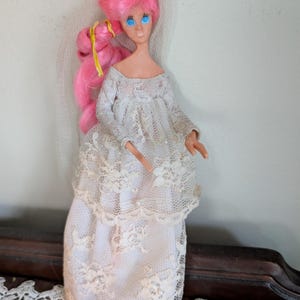 Puede incluir: Una muñeca vintage con cabello rosado y ojos azules, vestida con un vestido de novia de encaje blanco. La muñeca tiene un velo y un adorno decorativo para la cabeza. El vestido presenta intrincados detalles de encaje.