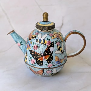 May include: A light blue teapot featuring a hand-painted pattern of various colorful butterflies, bees, and small pink flowers. The teapot has a brass-toned metal rim, handle, spout edge, and a small spherical knob on the lid with a bow detail.