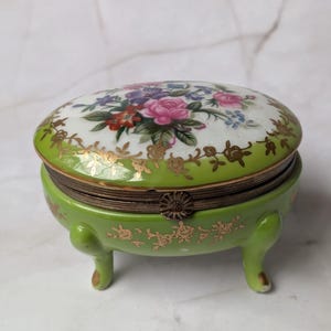 May include: An oval porcelain trinket box with a lime green base and lid, featuring gold floral accents and a central bouquet of pink, red, and blue flowers on the lid. The box stands on four small legs and has a metal hinge closure at the front.