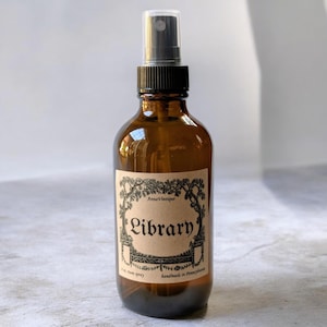 May include: An amber glass spray bottle with a black plastic nozzle and a tan paper label. The label features black decorative borders and the text AnnaVintique, Library, 4 oz. room spray, and handmade in Pennsylvania printed in black ink.