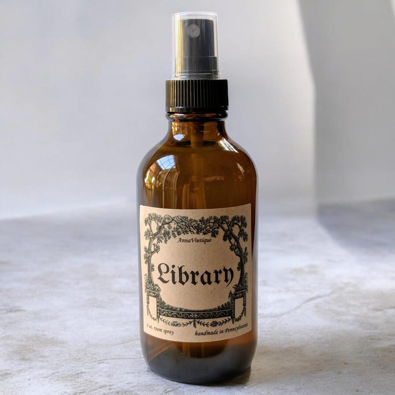 May include: An amber glass spray bottle with a black plastic nozzle and a tan paper label. The label features black decorative borders and the text AnnaVintique, Library, 4 oz. room spray, and handmade in Pennsylvania printed in black ink.