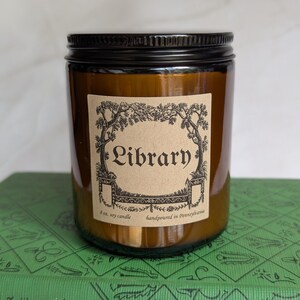 May include: An amber glass jar candle with a black metal lid and a tan paper label. The label features a black ink illustration of trees and the text Library, 8 oz. soy candle, handpoured in Pennsylvania. The candle sits on a green book.