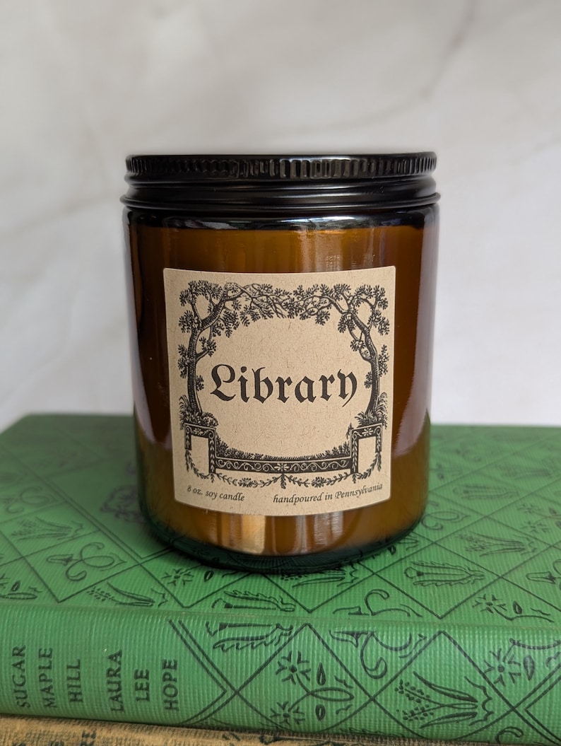 May include: An amber glass jar candle with a black metal lid and a tan paper label. The label features a black ink illustration of trees and the text Library, 8 oz. soy candle, handpoured in Pennsylvania. The candle sits on a green book.