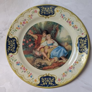 May include: A round decorative metal plate featuring a central illustration of two figures in a garden setting. The border is cream-colored with dark blue panels, gold accents, and a repeating pattern of small pink, yellow, and blue flowers.