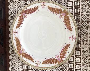Peut inclure: Une assiette plate en céramique blanche avec une bordure florale dorée et rose. L'assiette a un bord festonné et un rebord surélevé.