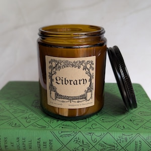 May include: An amber glass candle jar with a black metal lid, featuring a tan label with black text that reads Library, 8 oz. soy candle, handpoured in Pennsylvania, framed by a decorative illustration of trees and architectural elements.
