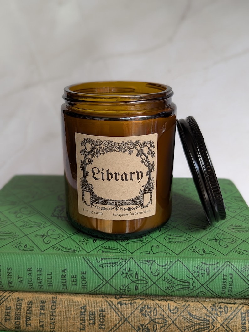 May include: An amber glass candle jar with a black metal lid, featuring a tan label with black text that reads Library, 8 oz. soy candle, handpoured in Pennsylvania, framed by a decorative illustration of trees and architectural elements.