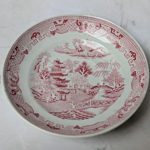 May include: A round ceramic plate featuring a detailed red and white transferware pattern. The design depicts a traditional landscape with pagodas, a bridge, trees, and water. The rim is decorated with a geometric and floral border pattern.