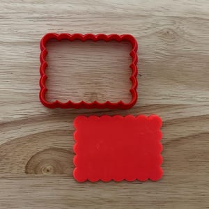 Puede incluir: Dos cortadores de galletas rectangulares rojos con bordes festoneados sobre una superficie de madera. Uno es un rectángulo completo, el otro un rectángulo sólido con un borde festoneado.