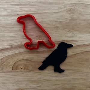 May include: A red plastic raven-shaped cookie cutter beside a black raven-shaped object. The cookie cutter is a simple outline of the bird. The raven-shaped object is solid black. Both are on a light brown wooden surface.