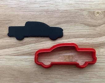 以下が含まれることがあります： 2つのトラック型のクッキーカッター。1つは黒いシルエットで、もう1つは赤いアウトラインです。どちらも明るい茶色の木製の表面に配置されています。カッターは、トラック型のクッキーやお菓子を作るように設計されています。